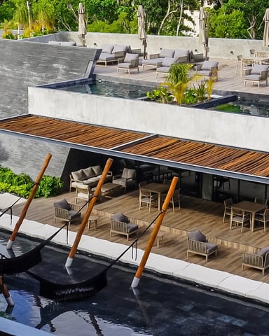 Modern resort pool deck with wooden loungers, shaded pavilion, multiple pools, and lush tropical landscaping
