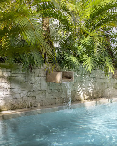 Swimming pool with turquoise water and waterfall feature, stone wall, and lush palm trees overhead